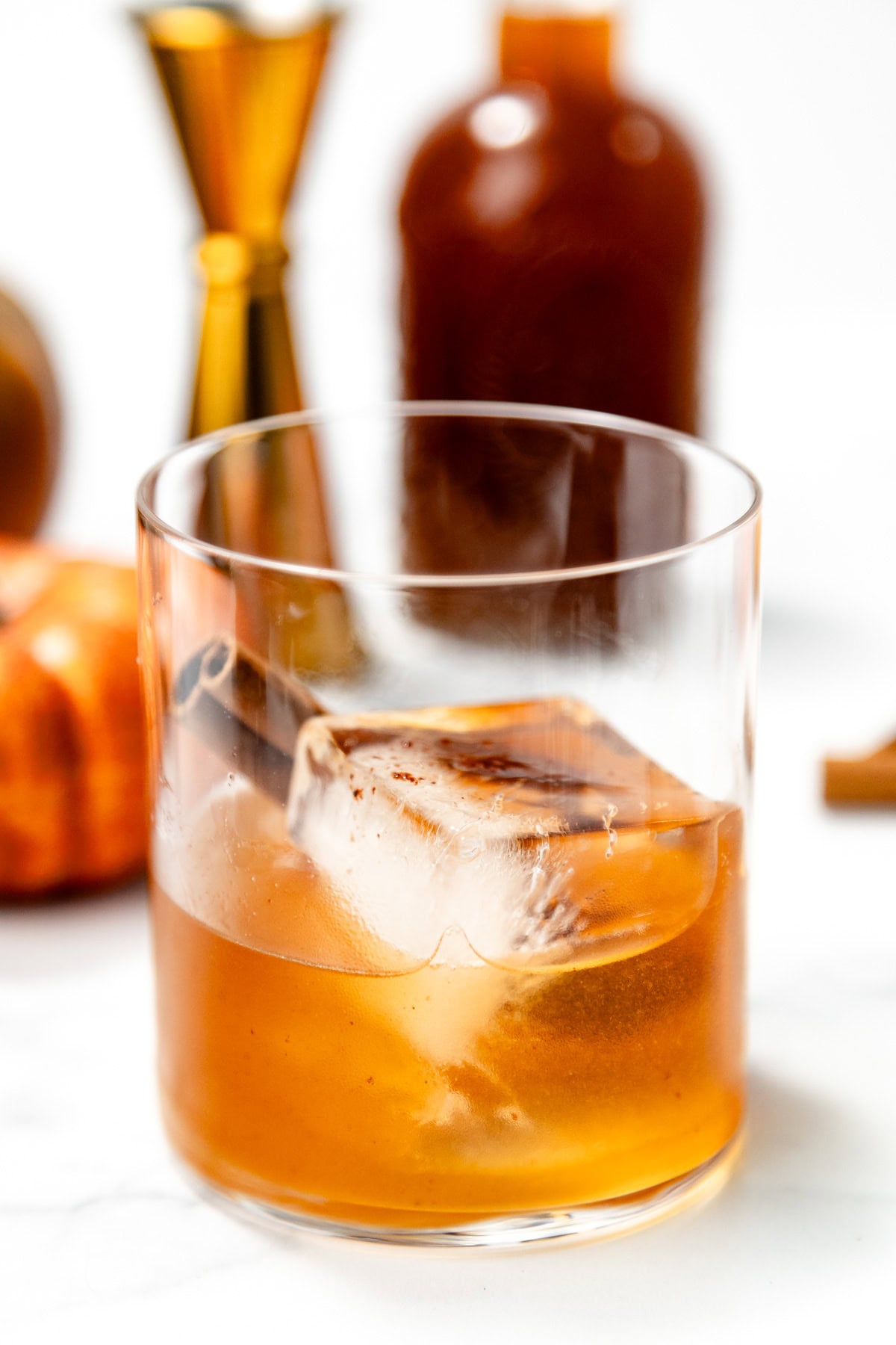 A glass of amber cocktail with a large ice cube and cinnamon stick, with bottles and pumpkin in the background.