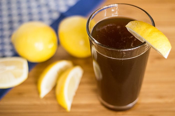 A glass of dark liquid with a lemon wedge, next to whole and sliced lemons on a wooden surface.