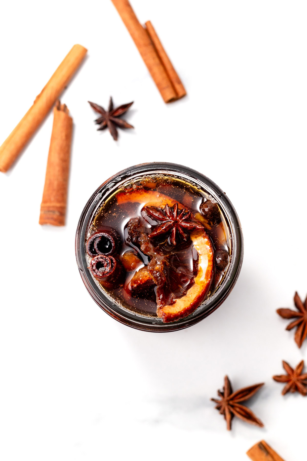 Top view of a glass with spiced drink, orange slices, star anise, and cinnamon sticks on a white surface.