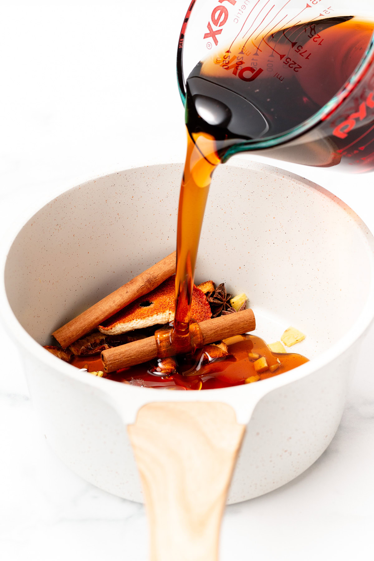 Maple syrup is poured into a white saucepan with cinnamon sticks, ginger, and star anise inside.