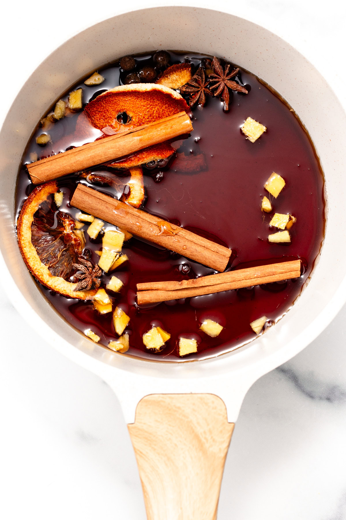 A saucepan with mulled wine, orange slices, cinnamon sticks, ginger pieces, and spices on a white surface.