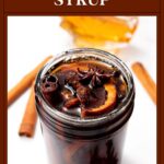 A jar of spiced maple syrup with orange slices, cinnamon sticks, and star anise on a white background.
