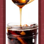 A spoon drizzles spiced maple syrup into a glass jar filled with orange peels and spices.