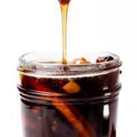 Syrup being poured from a ladle into a jar filled with citrus peels and dark liquid.