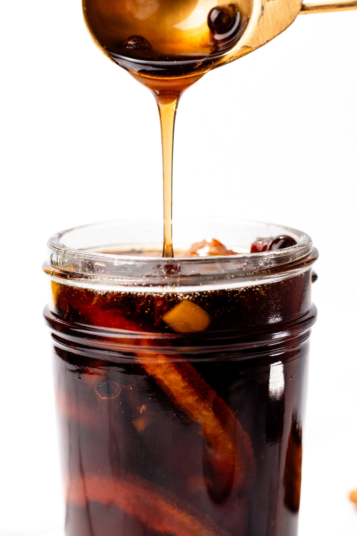 Syrup being poured from a ladle into a jar filled with citrus peels and dark liquid.