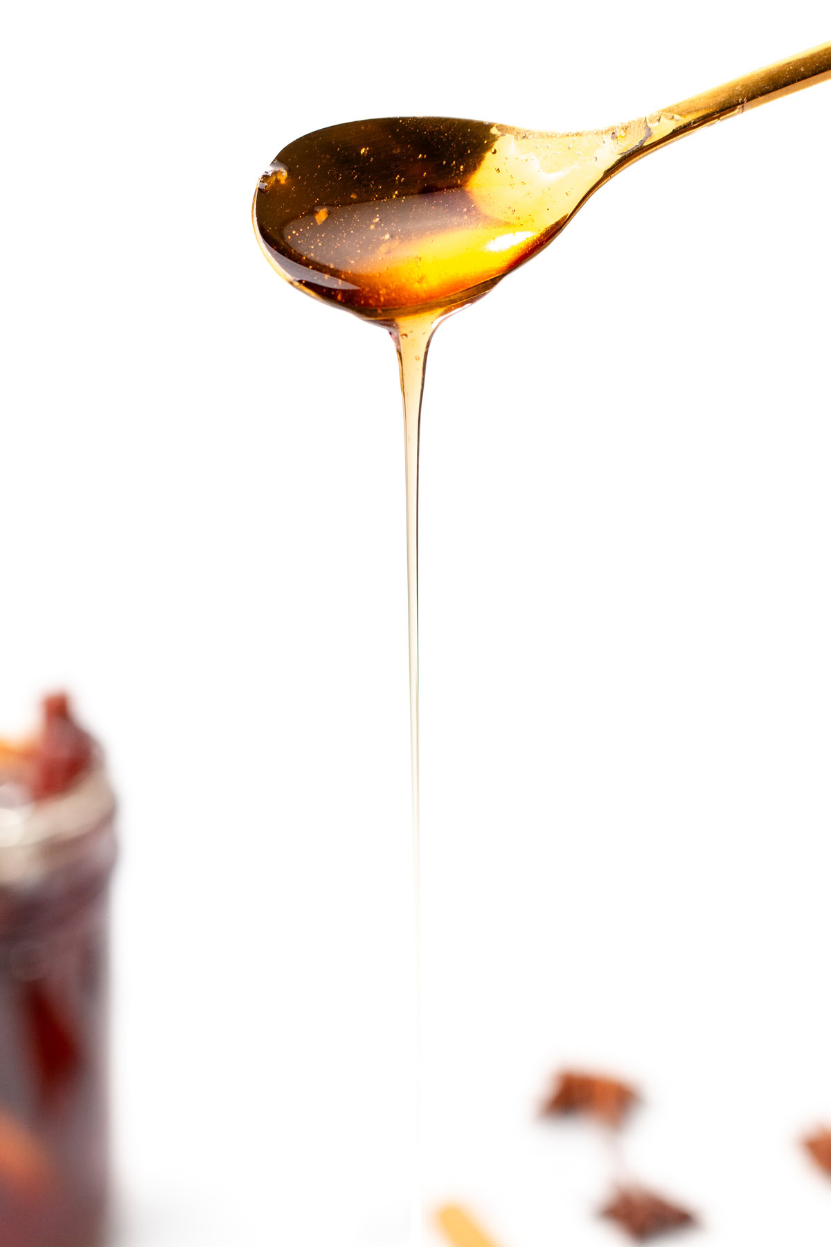 A spoon drizzling golden honey, with a blurred jar and spices in the background on a white surface.
