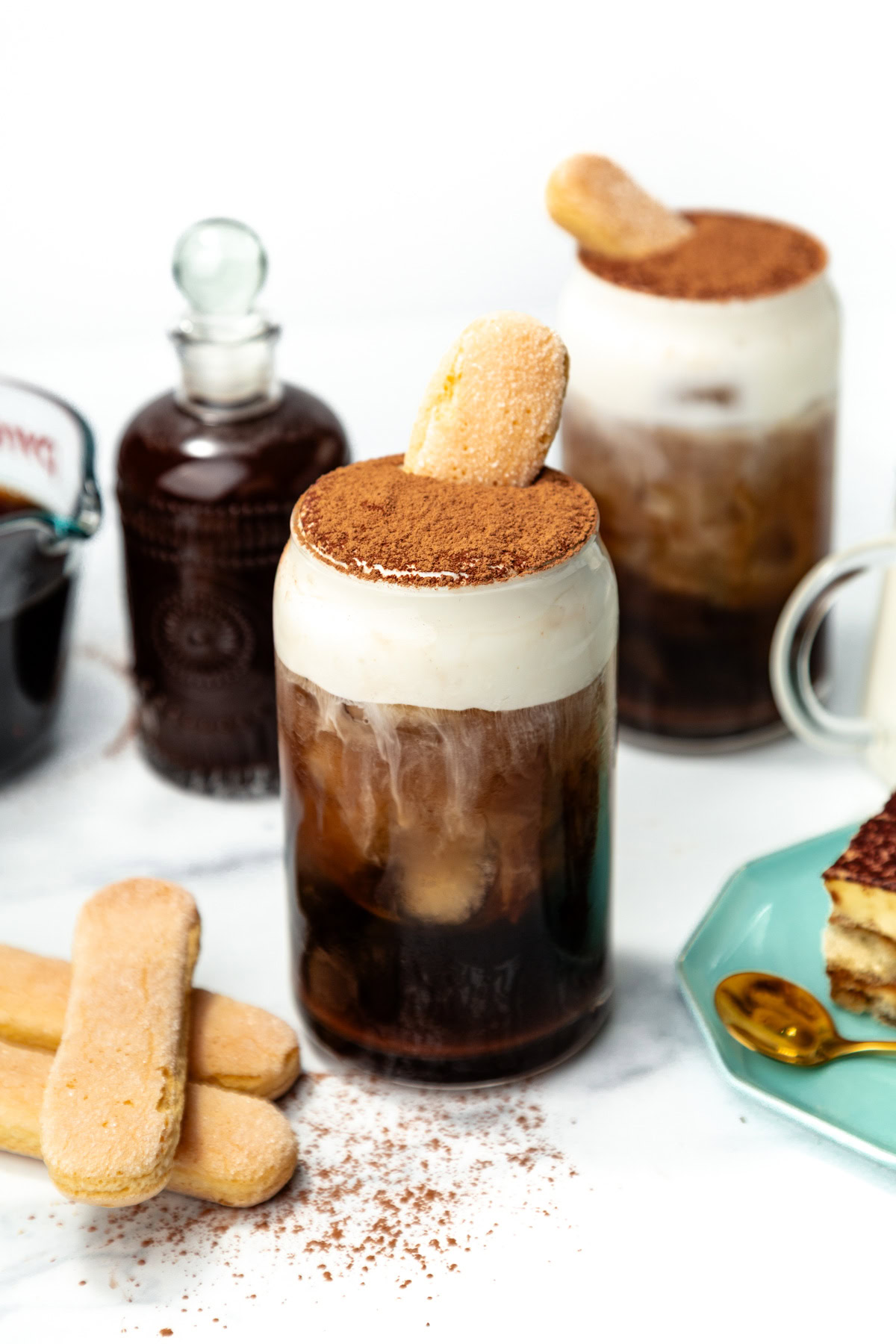 Iced coffee with foam, dusted cocoa, and a ladyfinger biscuit in a glass, with tiramisu and biscuits nearby.