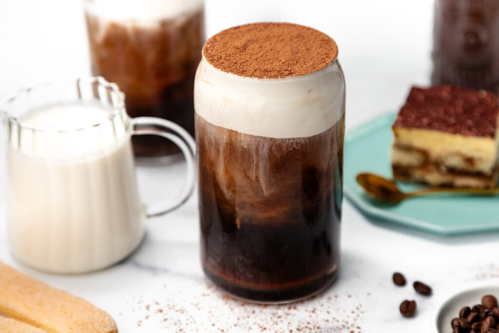 A glass of iced coffee topped with cream and cocoa powder, next to milk and a slice of tiramisu.