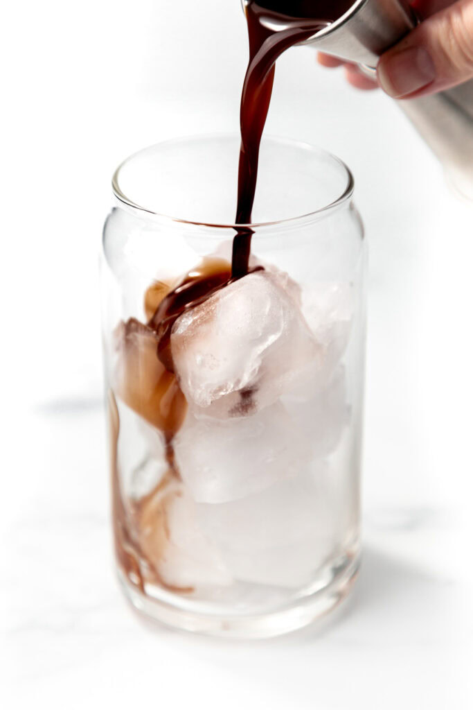 Dark liquid being poured over ice cubes in a clear glass, with a hand holding a metal cup.