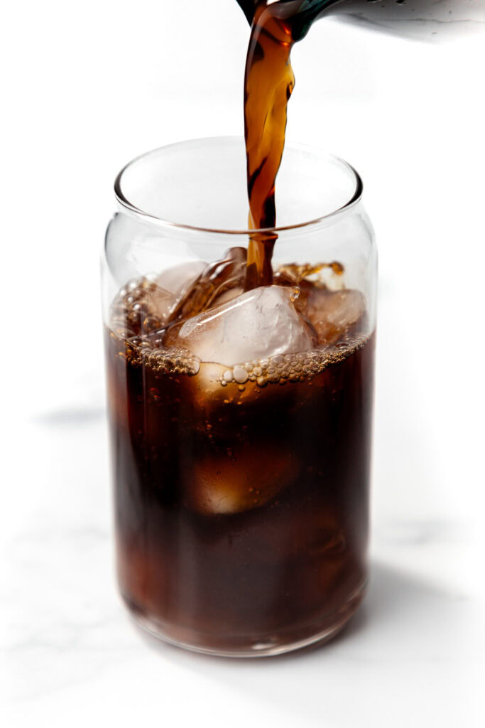 Cola being poured over ice cubes in a glass on a white background.