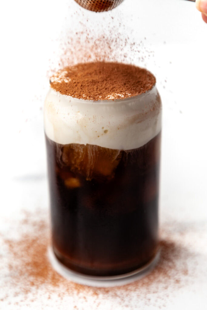 Iced coffee in a glass with foam on top, being dusted with cocoa powder against a white background.