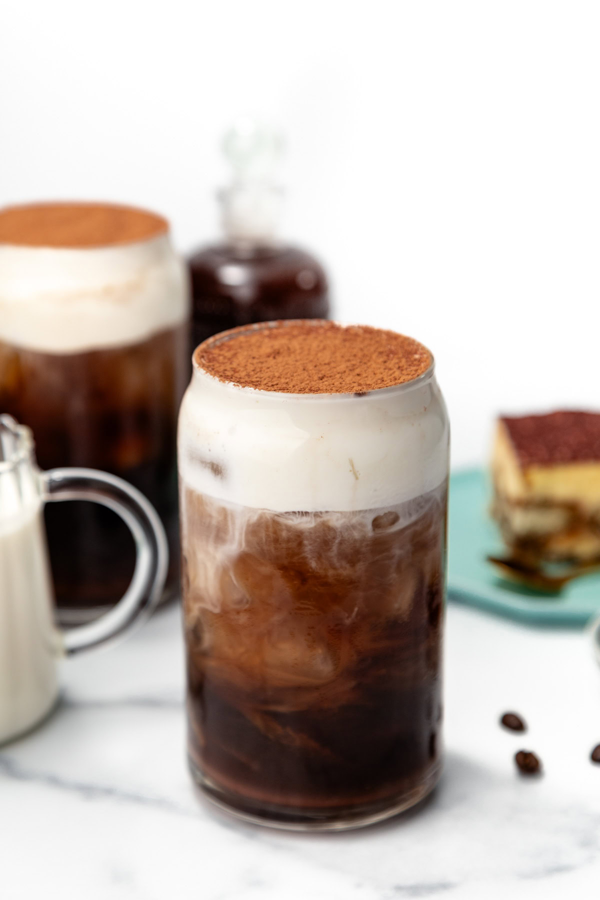 Iced coffee in a glass with creamy foam and cocoa powder on top, surrounded by coffee and dessert.