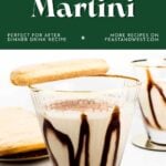 A tiramisu martini in a glass with chocolate drizzle, surrounded by ladyfingers on a white surface.