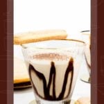 A tiramisu martini in a glass with chocolate drizzle, surrounded by ladyfingers on a white surface.