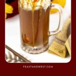 A glass mug filled with a frothy drink, topped with caramel, sits beside chocolate and a scarf.