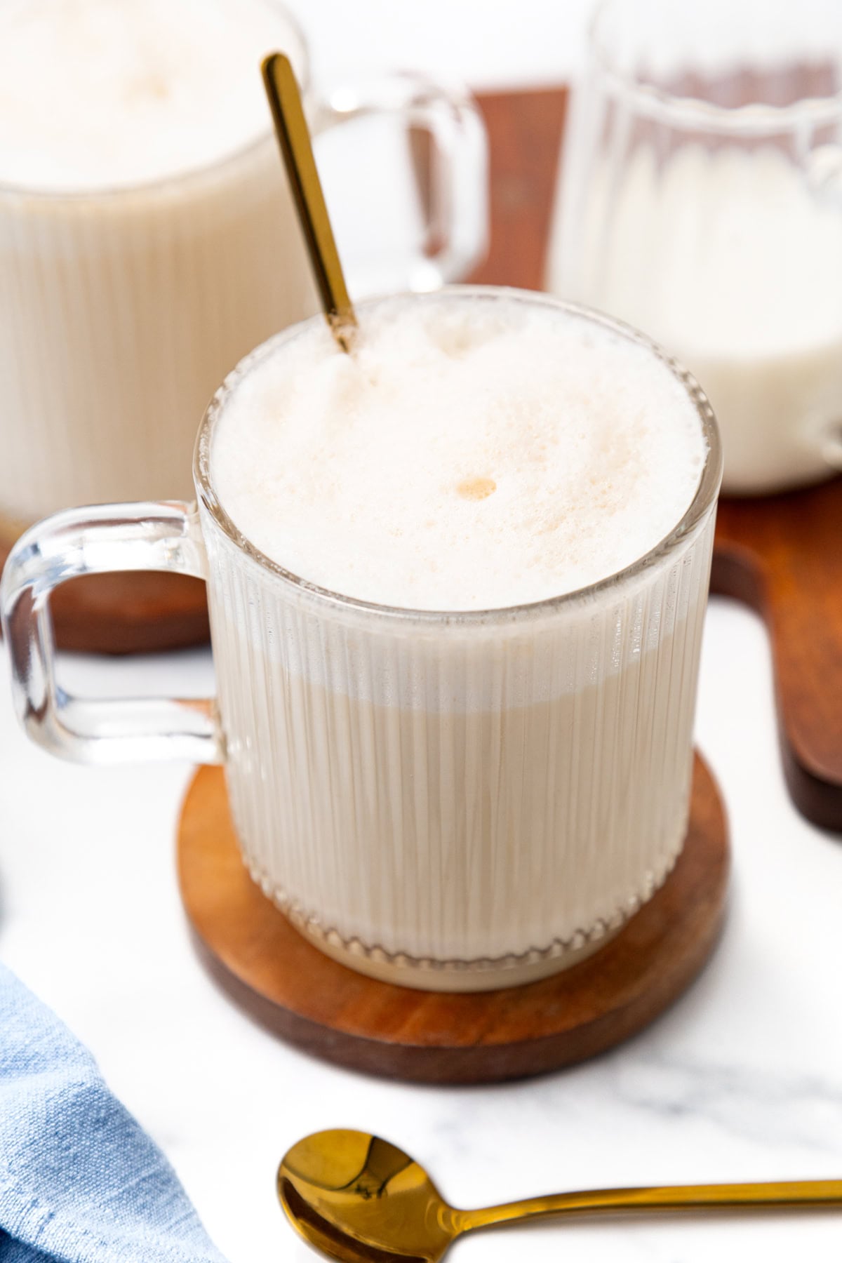 A glass mug filled with a foamy latte, resting on a wooden coaster with a gold spoon.