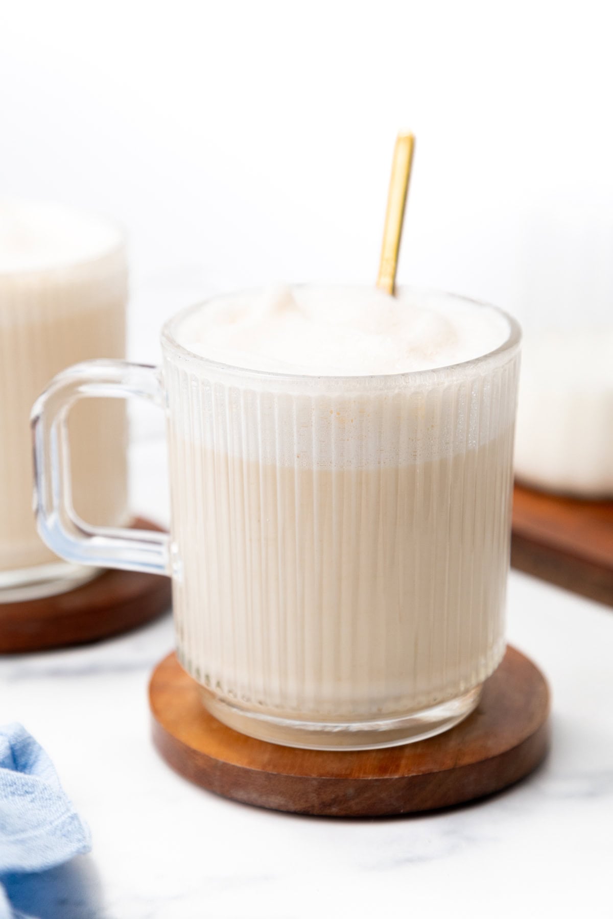 A glass mug of latte with a gold spoon, resting on a wooden coaster.