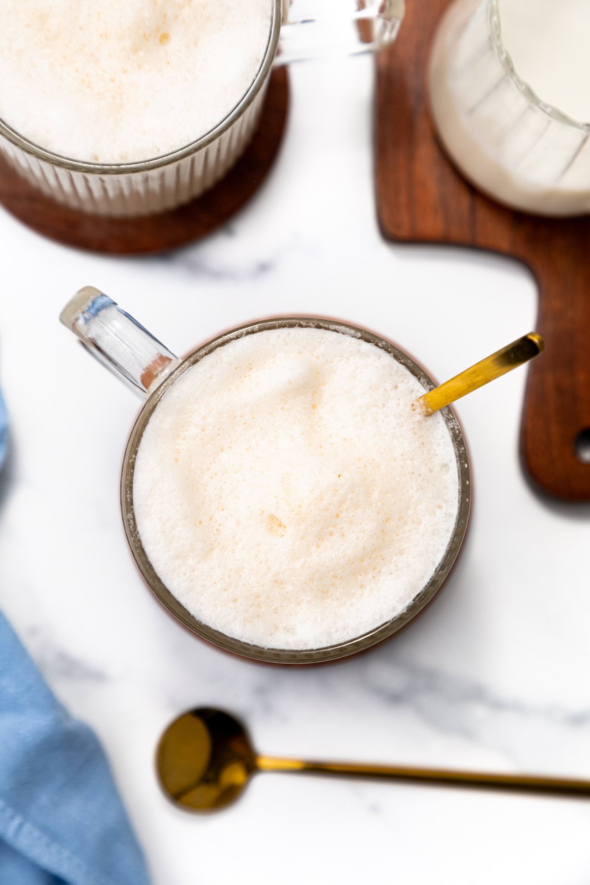 Top-down view of two glass mugs filled with frothy coffee, with gold spoons and a small pitcher of milk nearby.
