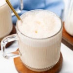 A glass mug of foamy latte with a gold spoon on a wooden coaster.