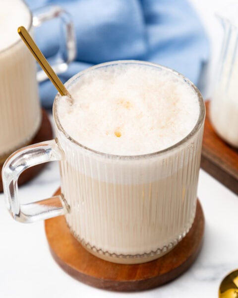 A glass mug of foamy latte with a gold spoon on a wooden coaster.