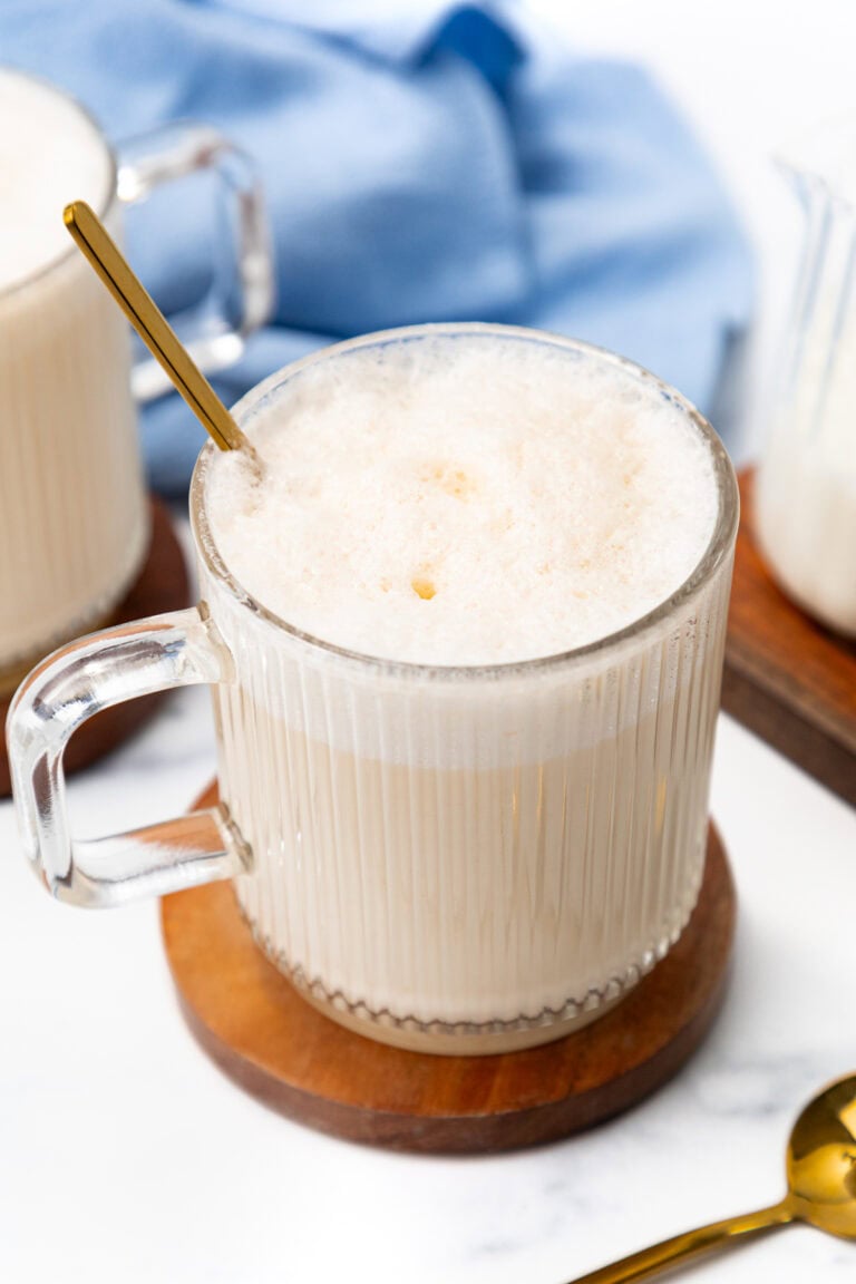 A glass mug of foamy latte with a gold spoon on a wooden coaster.