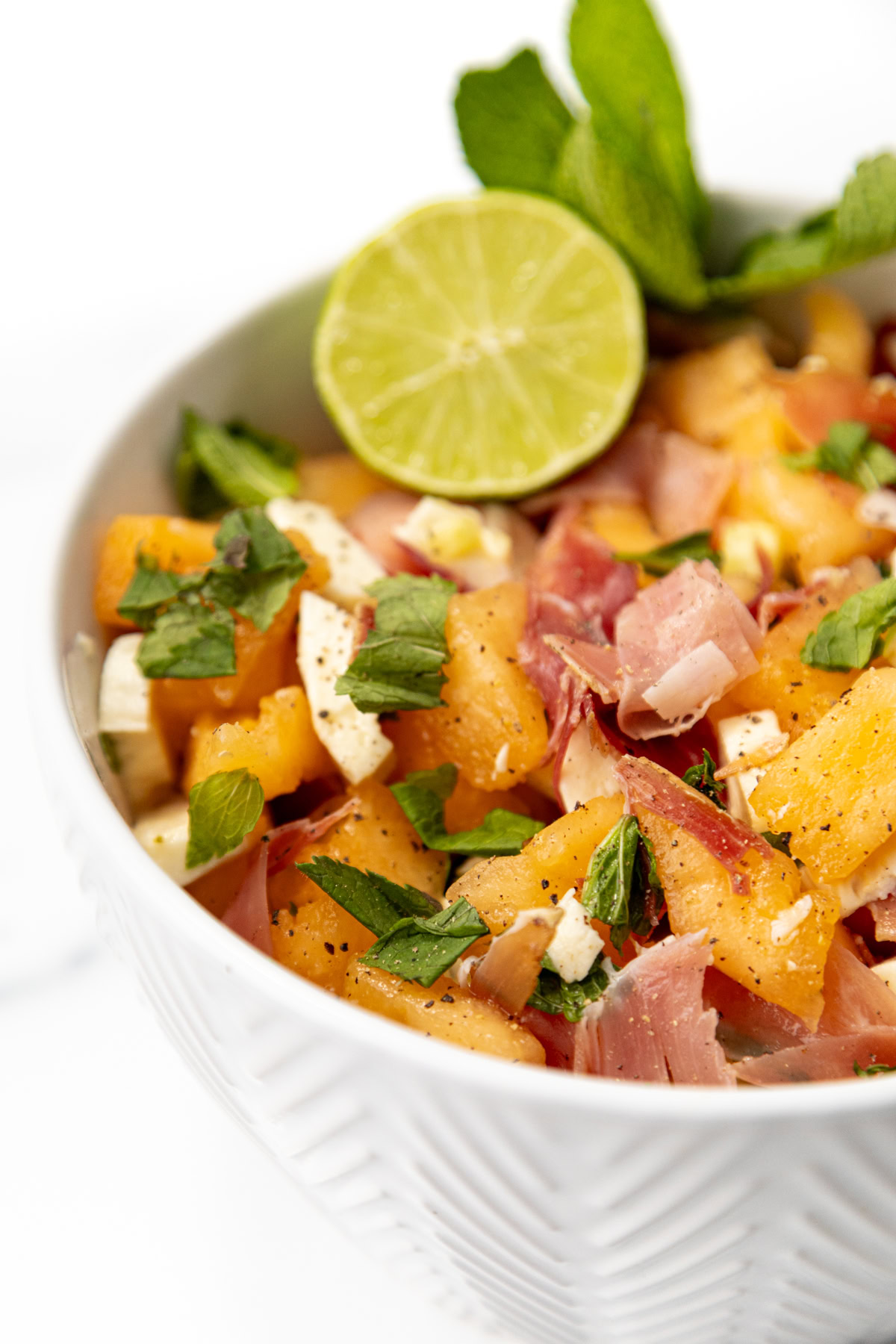 Bowl of cantaloupe, prosciutto, and mozzarella salad topped with a lime slice and fresh mint.