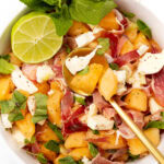 Bowl of melon, prosciutto, and mozzarella salad garnished with mint and a lime wedge.