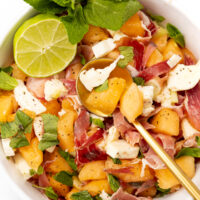 Bowl of melon, prosciutto, and mozzarella salad garnished with mint and a lime wedge.
