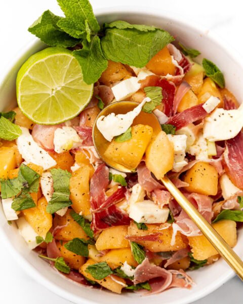 Bowl of melon, prosciutto, and mozzarella salad garnished with mint and a lime wedge.