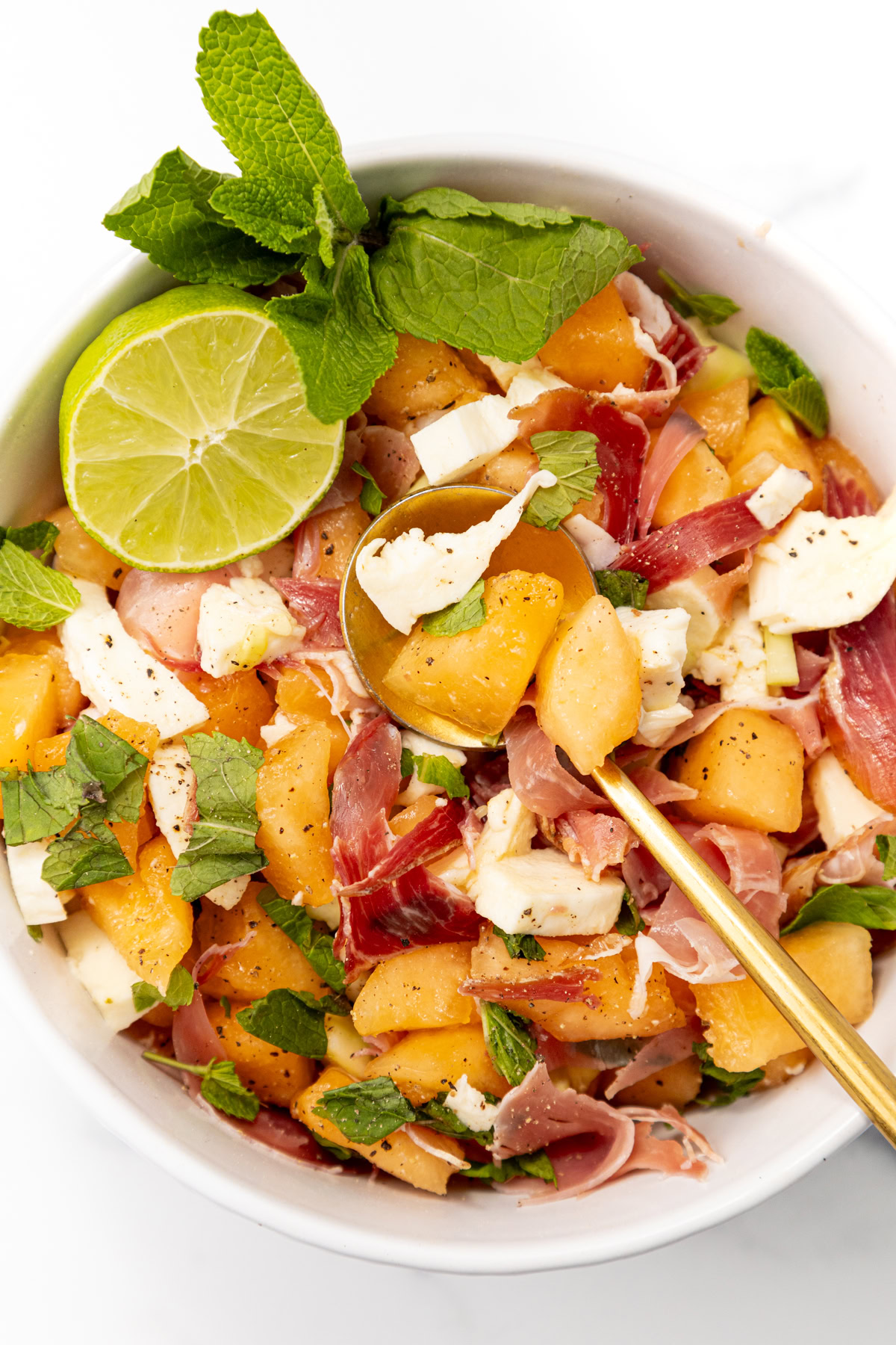 Bowl of melon, prosciutto, and mozzarella salad garnished with mint and a lime wedge.