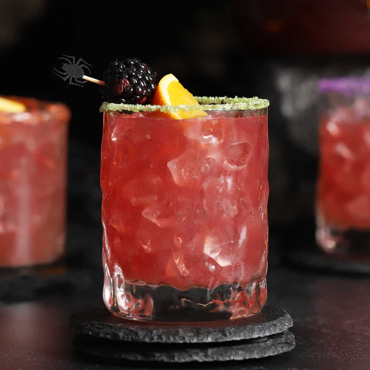 Red cocktail with ice, garnished with a blackberry, orange slice, and a decorative spider pick.
