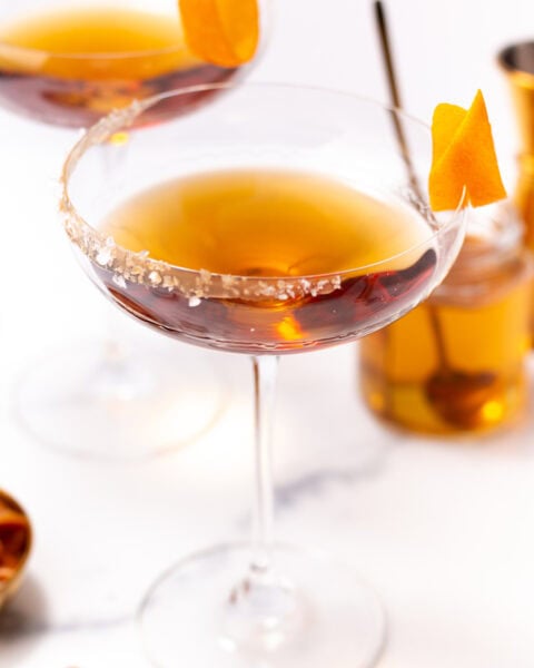Two cocktail coupes filled with amber liquid and garnished with orange twists and salted rims.