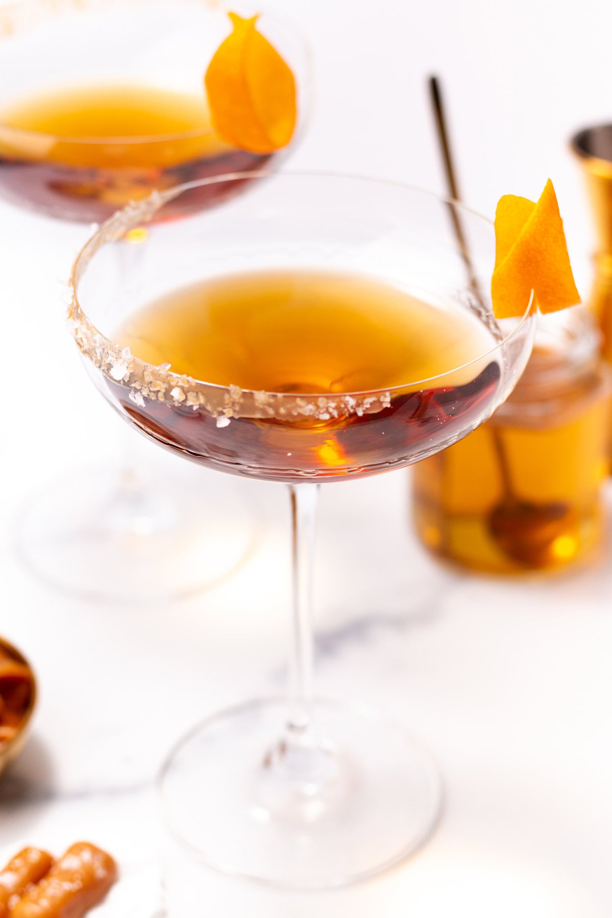 Two cocktail coupes filled with amber liquid and garnished with orange twists and salted rims.