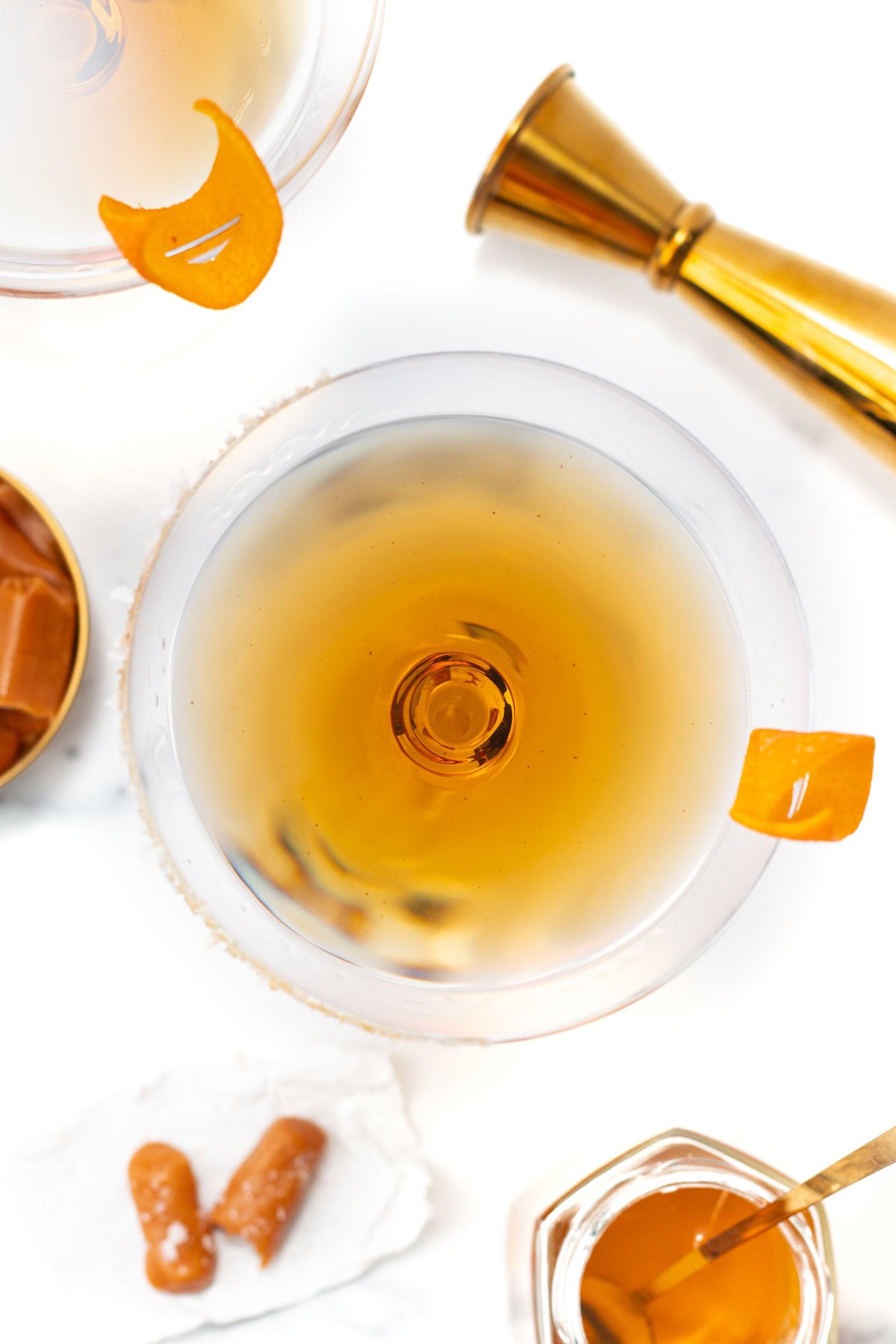 Top-down view of two cocktails garnished with orange peel, next to a jigger and caramel candies.