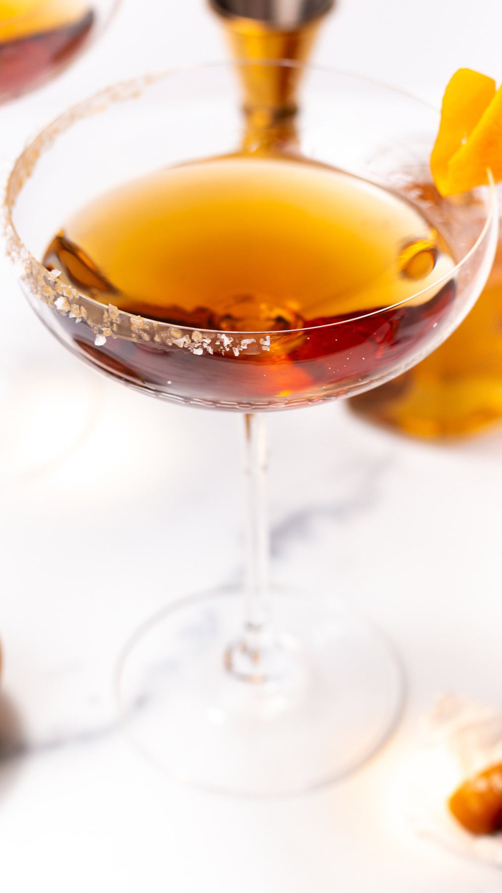 A cocktail in a stemmed glass with a salt-rimmed edge and an orange twist garnish.