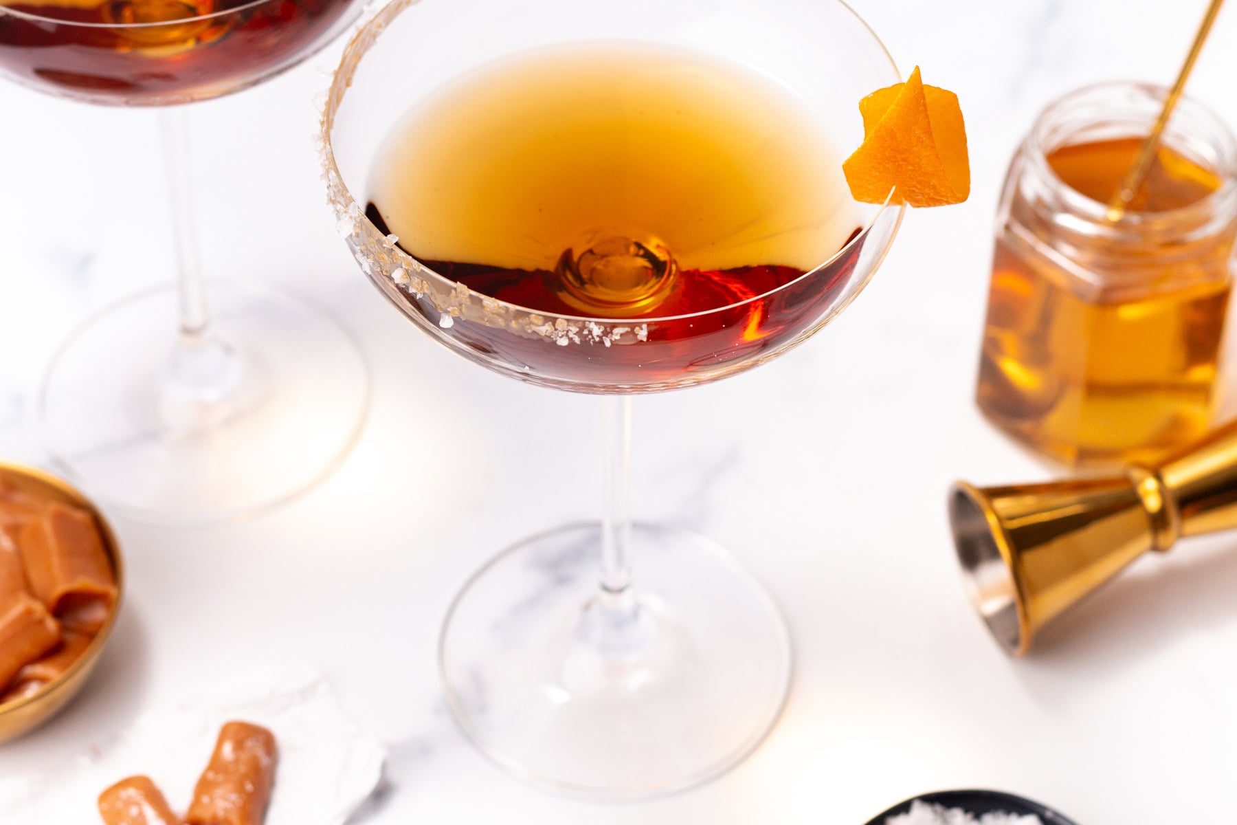 Cocktail in a rimmed coupe glass garnished with an orange twist, next to a jar of honey and a jigger.