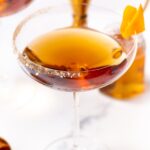 A coupe glass filled with an amber cocktail, rimmed with salt and garnished with an orange twist.