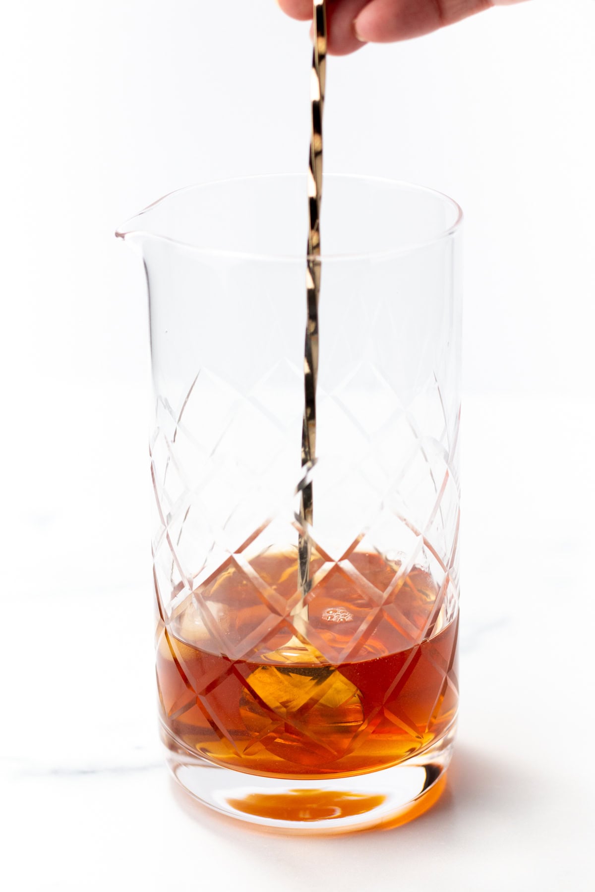 A hand uses a long bar spoon to stir an amber cocktail in a crystal mixing glass.