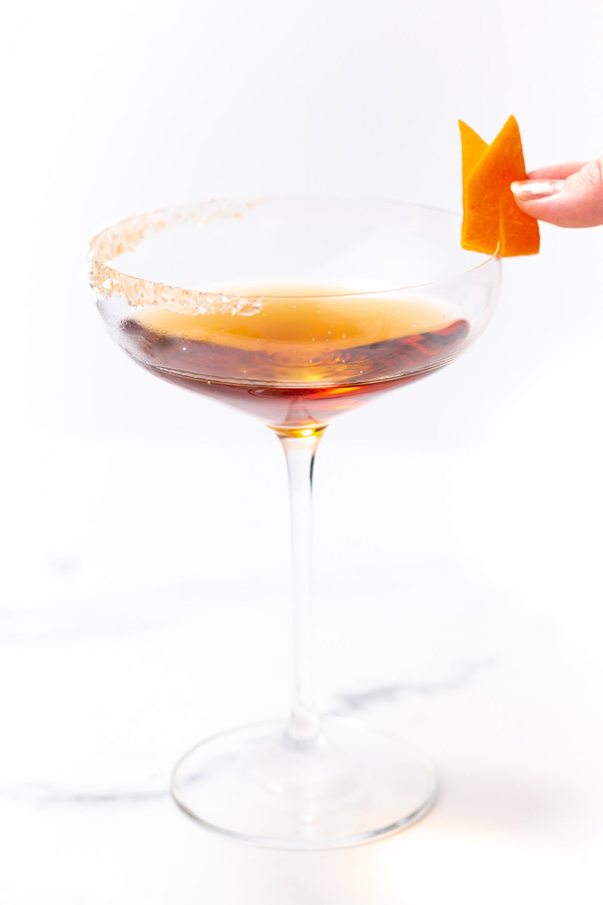 A hand garnishes a sugar-rimmed coupe glass filled with an amber cocktail with an orange twist.