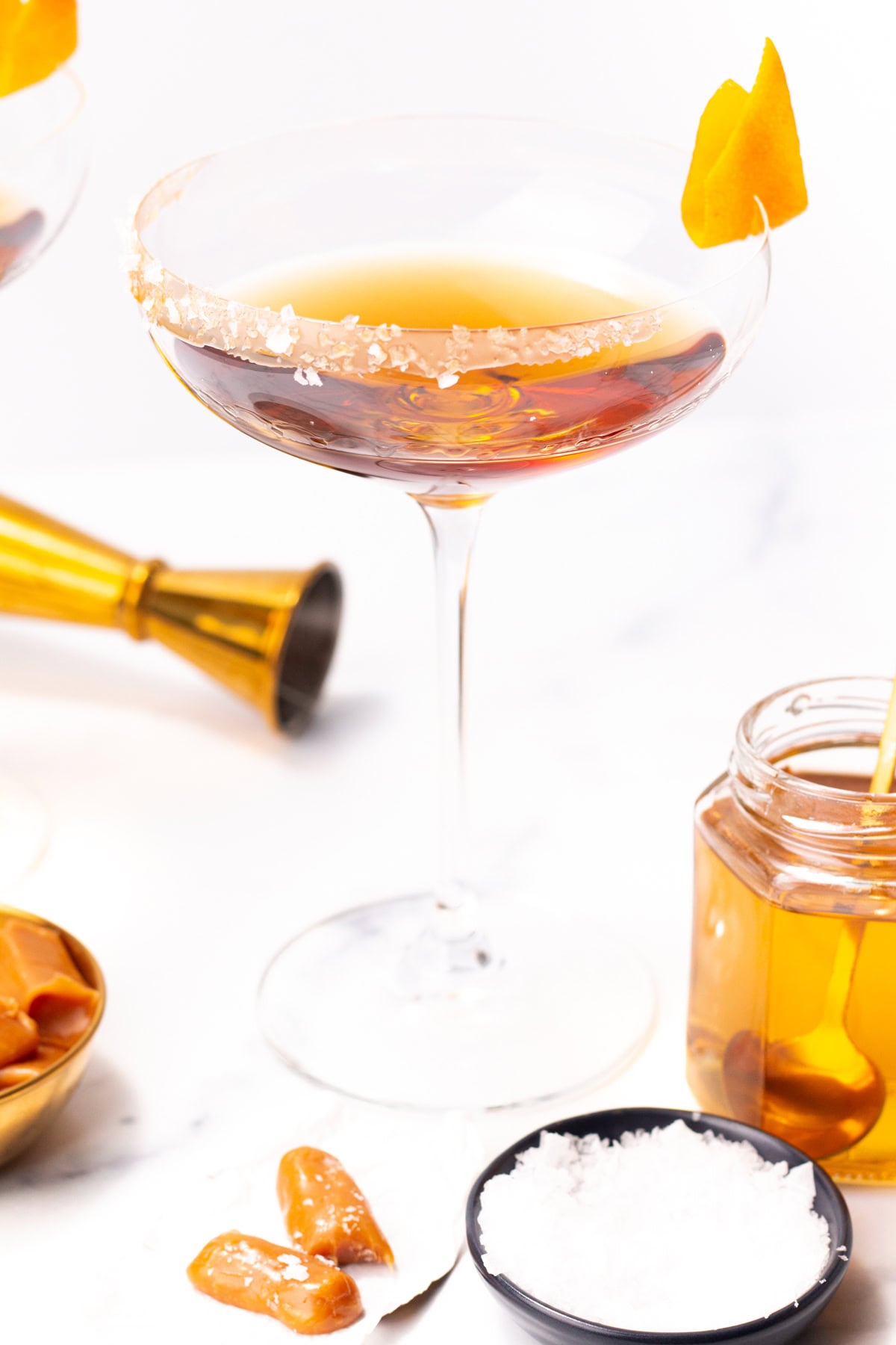 A cocktail in a salt-rimmed coupe glass with an orange twist, alongside honey and salted caramels.