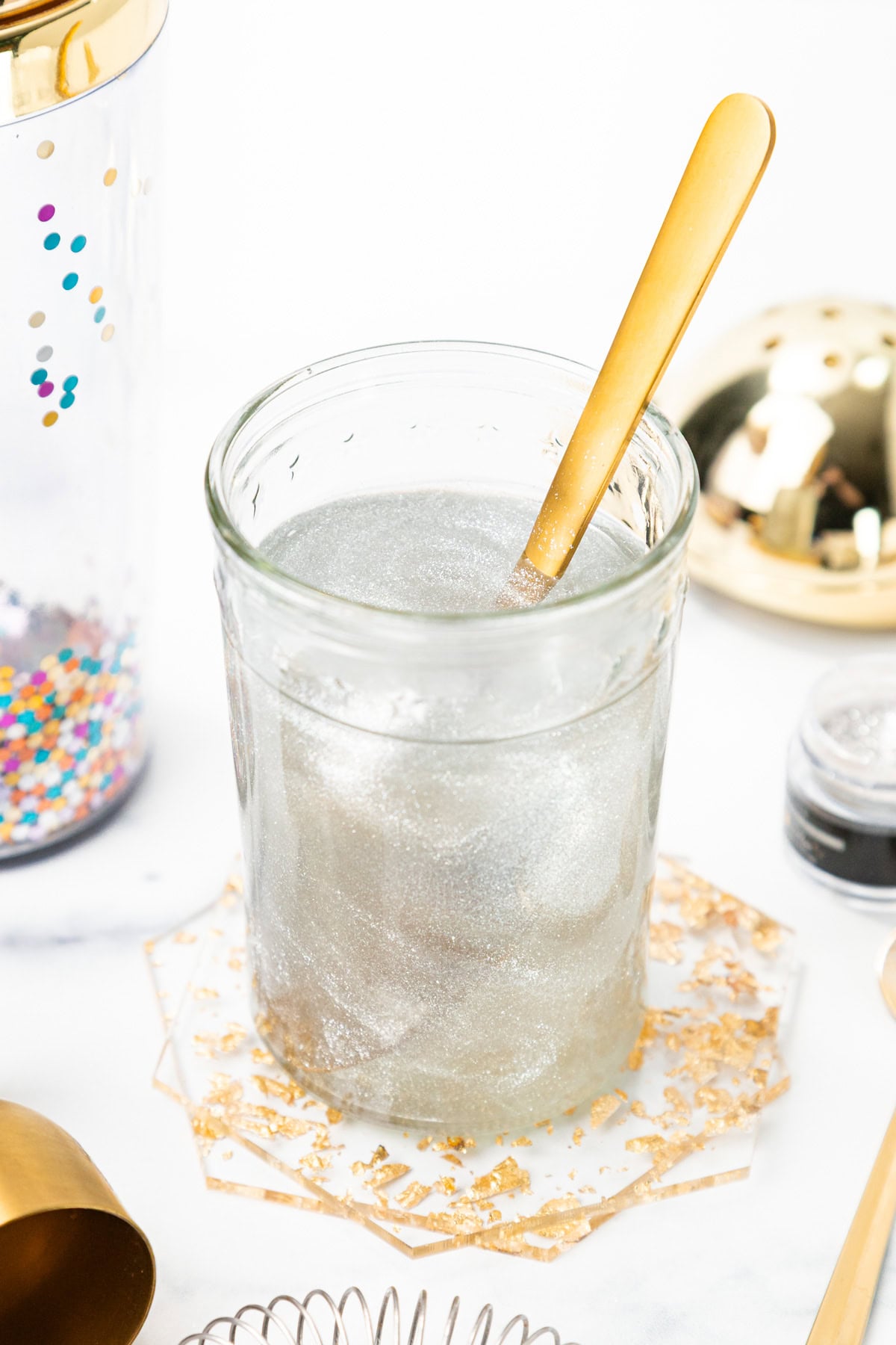 Glass of silver glittery liquid with a gold spoon, sitting on a gold-flecked coaster.