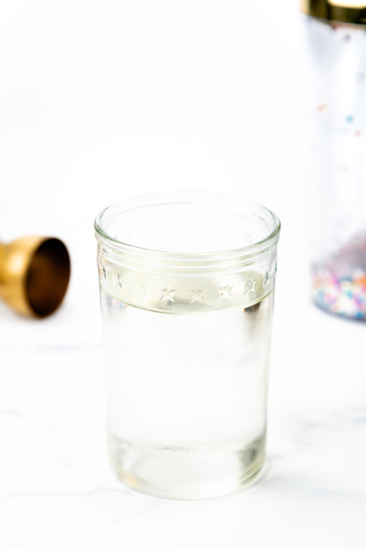A glass of clear liquid with a star-patterned rim, beside a gold jigger and a shaker.