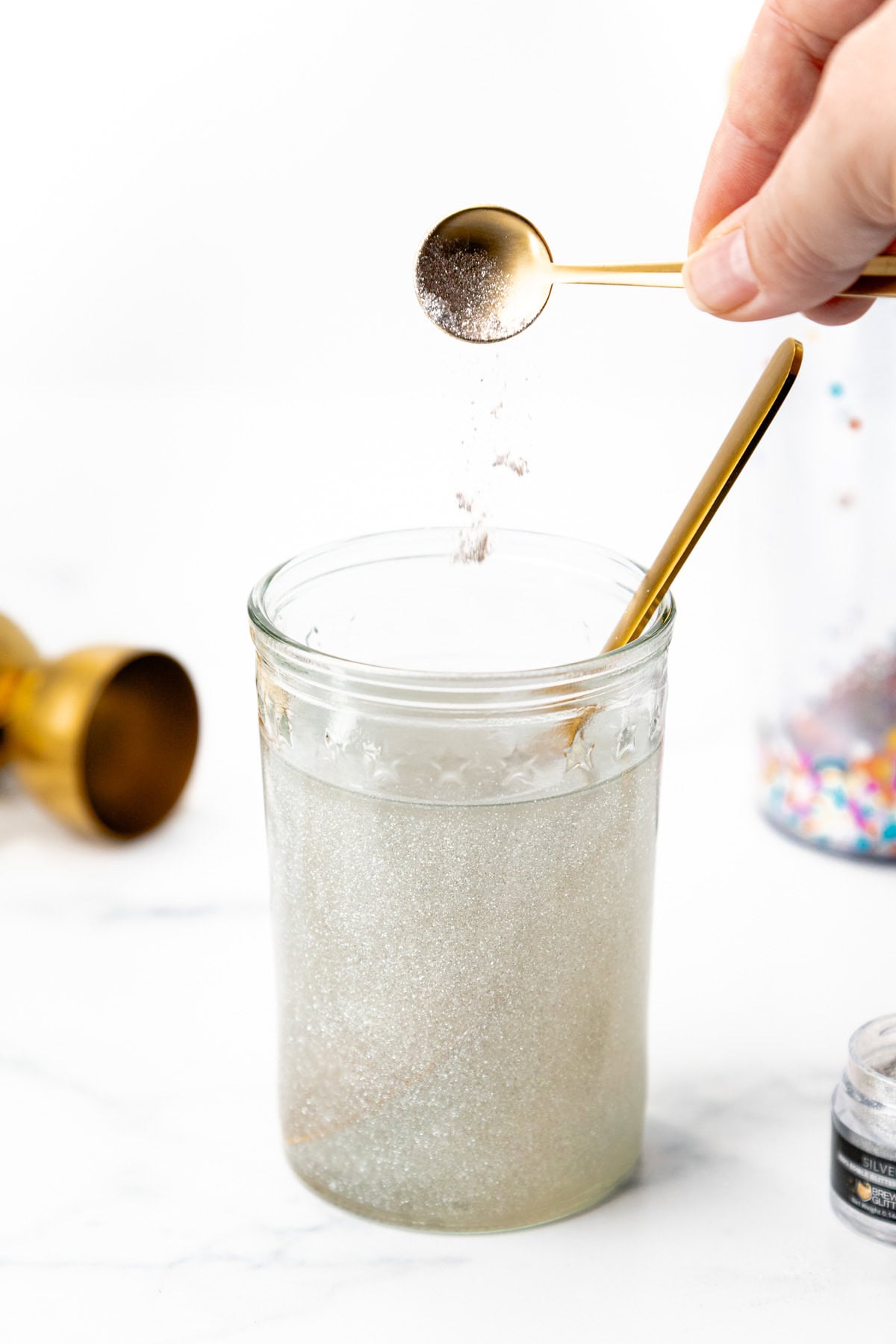 A hand uses a small gold spoon to sprinkle edible silver glitter into a glass of liquid.