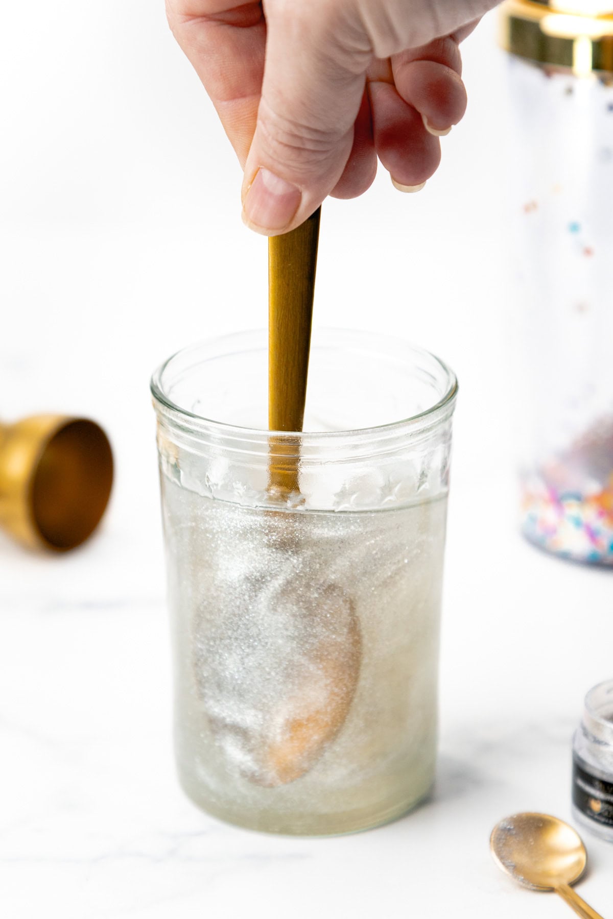 A hand stirs a shimmery silver liquid in a glass jar with a wooden spoon.