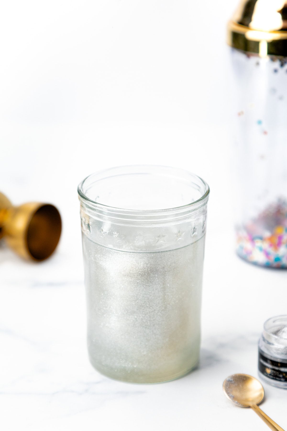 A glass filled with shimmering silver liquid, with a cocktail shaker and a small jar of glitter nearby.