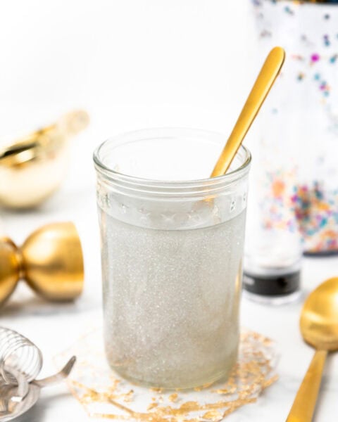 A glass of shimmering silver cocktail with a gold spoon, surrounded by bar tools.