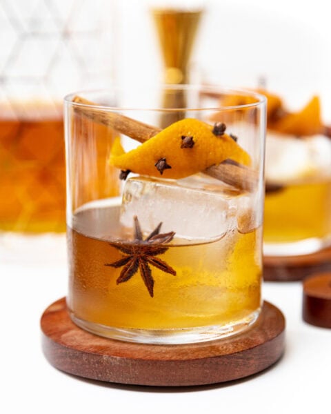 Cocktail on a wooden coaster garnished with a star anise, cinnamon stick, and orange peel studded with cloves.