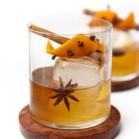 Cocktail in a glass with a large ice cube, star anise, cinnamon stick, and orange peel studded with cloves.