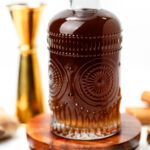A decorative glass bottle filled with dark liquid sitting on a wooden coaster, surrounded by spices.
