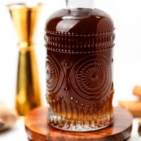 A decorative glass bottle filled with dark liquid sitting on a wooden coaster, surrounded by spices.